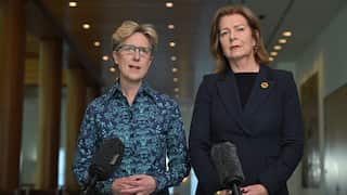 Two middle-aged women stand in front of microphones, one dressed in a patterned blue shirt and the other in a dark blazer.