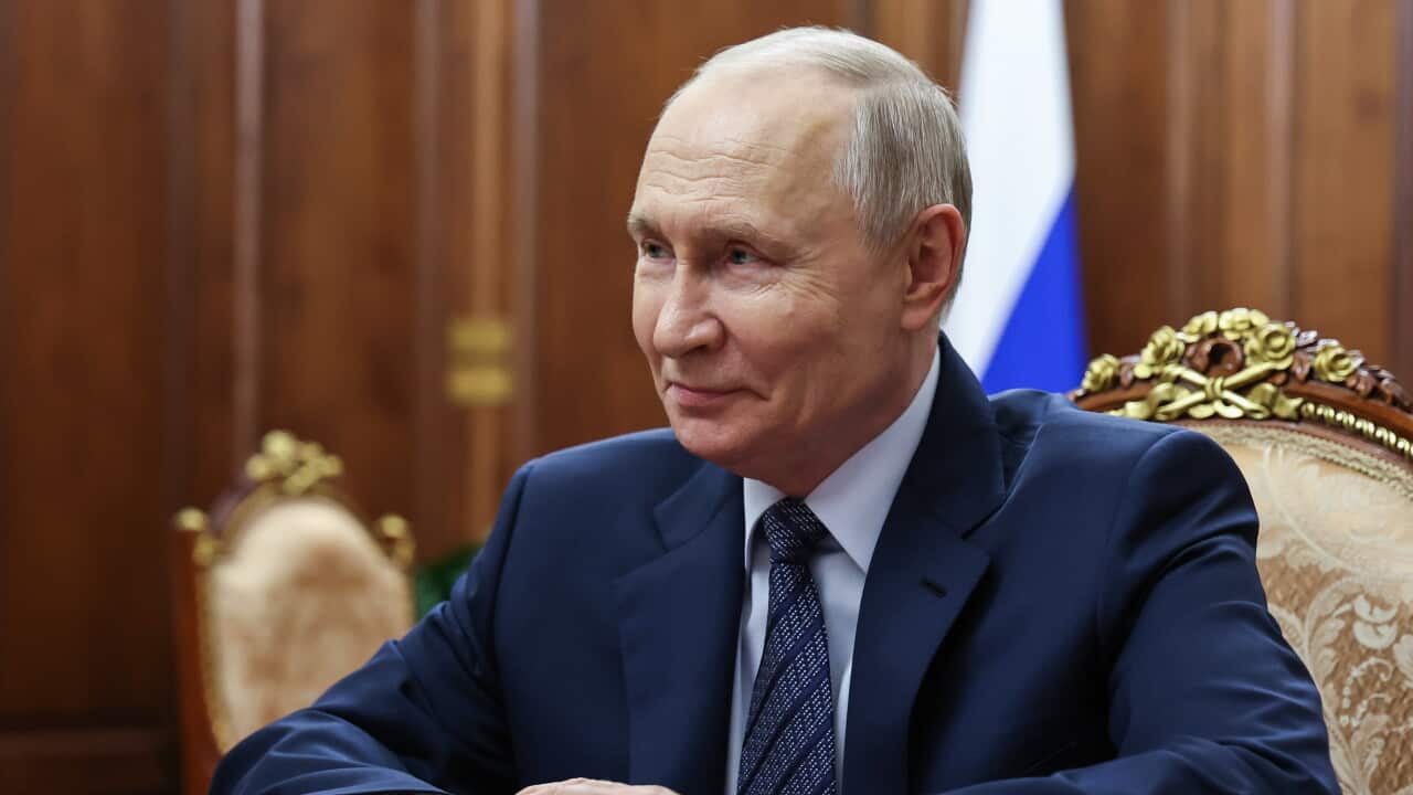 Man in a dark suit and tie sits at an ornate table in a wood-panelled room, with a Russian flag visible in the background.