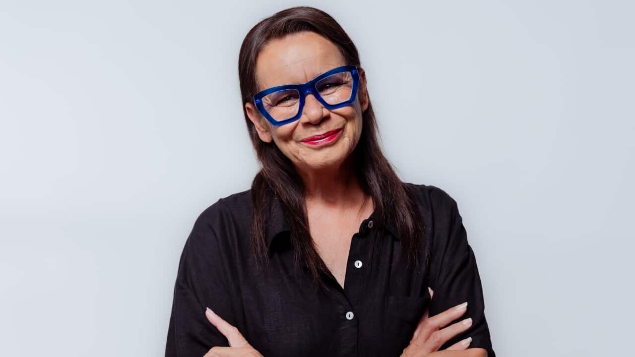 A smiling middle-aged woman with long dark hair stands against a plain light background.