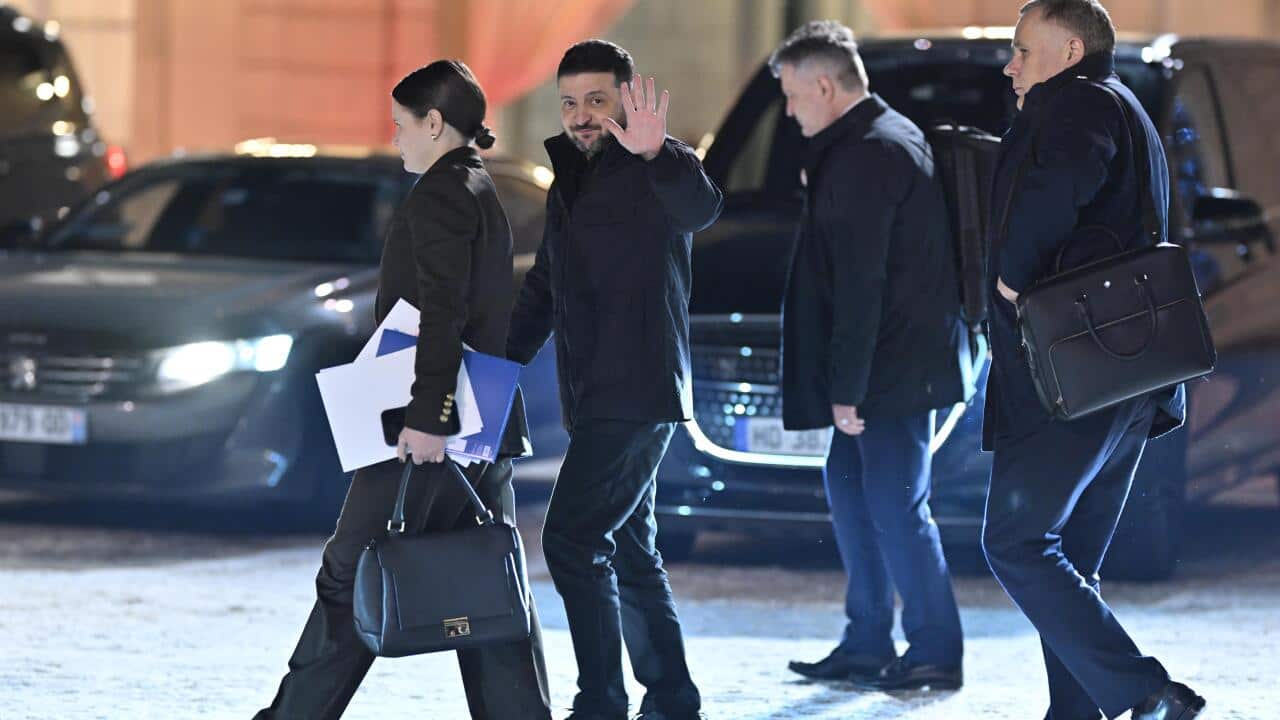 Ukrainian President Volodymyr Zelenskyy waves to the camera while walking outdoors with several staff members near parked vehicles.