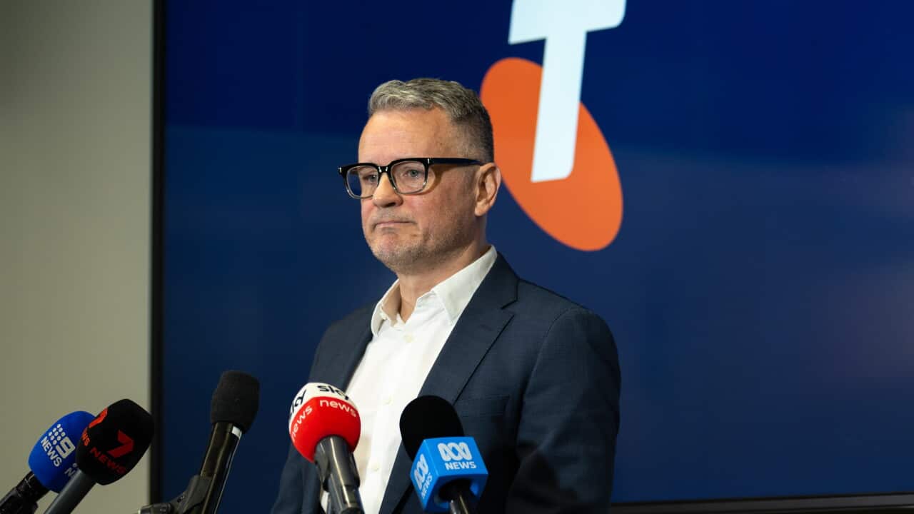 A middle aged white man in glasses and a suit stands in front of microphones and a Telstra logo.
