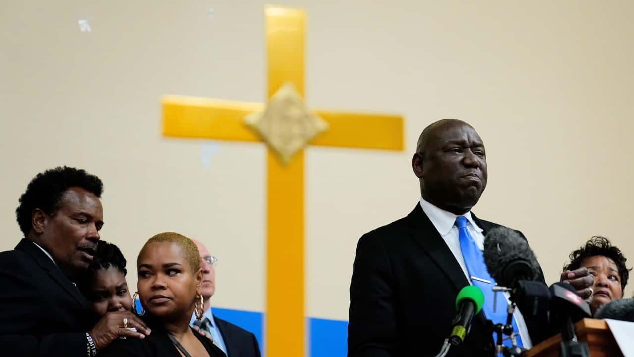 Lawyer Benjamin Crump stands at a podium speaking to reporters. A large cross is on the wall in the background.