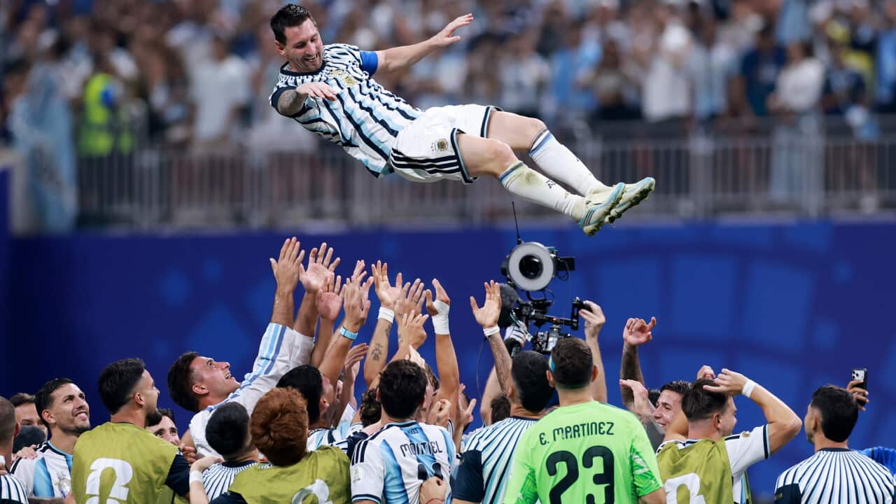 Lionel Messi in a white and blue uniform being lifted up in the air by his teammates on a football pitch.
