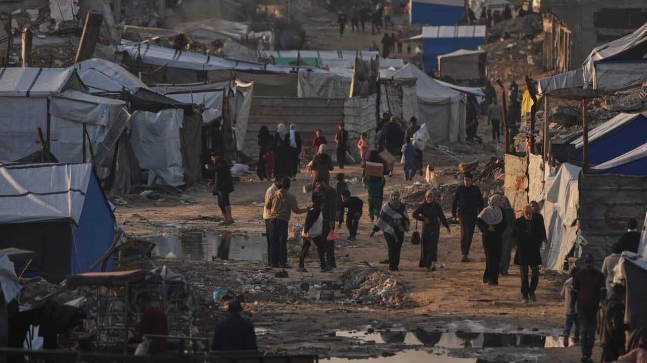 A crowded dirt path winds through a dense displacement camp of makeshift tents and crumbling concrete ruins as people navigate past puddles and debris.
