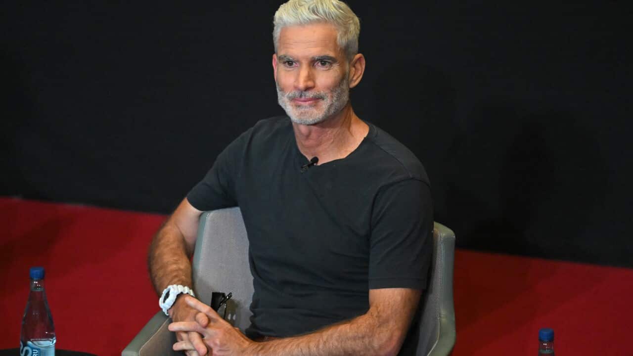 A man in a black shirt sitting on a chair positioned on a stage with a bottle of water next to him.