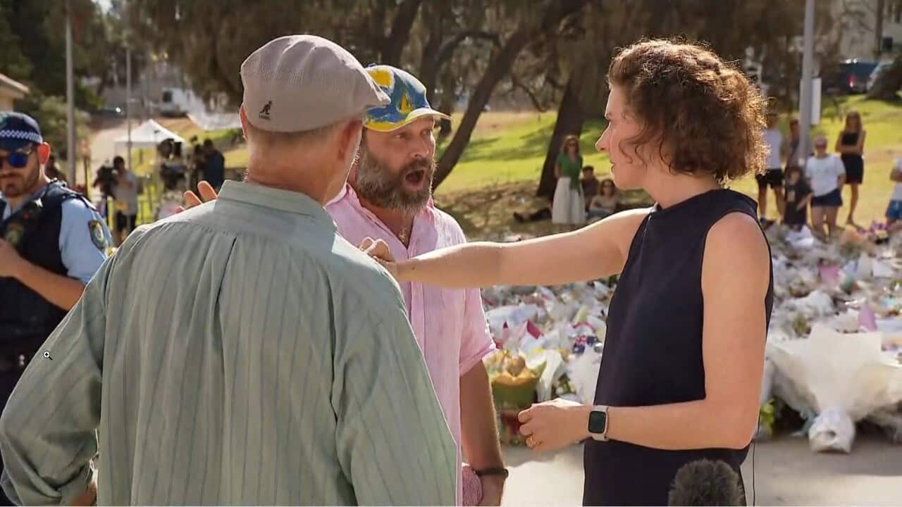 Allegra Spender holds her arm out as a man speaks animatedly with his mouth wide open. They are standing in front of a memorial area covered in flowers.