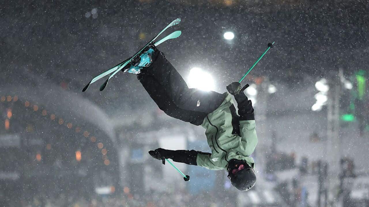 A person wearing a teal jacket and black pants flipping upside down in skis whilst it's snowing