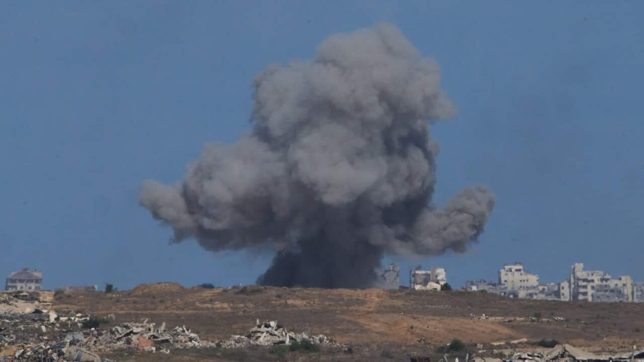 Black plumes of smoke billow over buildings at a distance following bombardment.