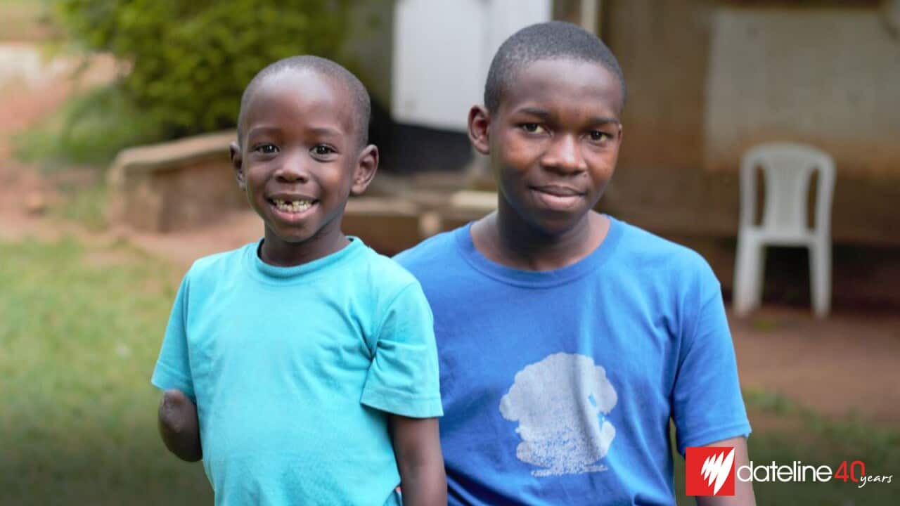 Two African boys wearing blue t-shirts are posing for a photo. The boy on the right is in his late teens; the boy on the left is seven years old and is missing a right arm