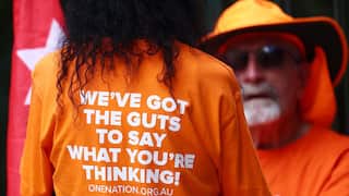 One Nation members in orange shirts. A woman's back is towards the camera, with her shirt saying 'We've got the gut to say what you're thinking'
