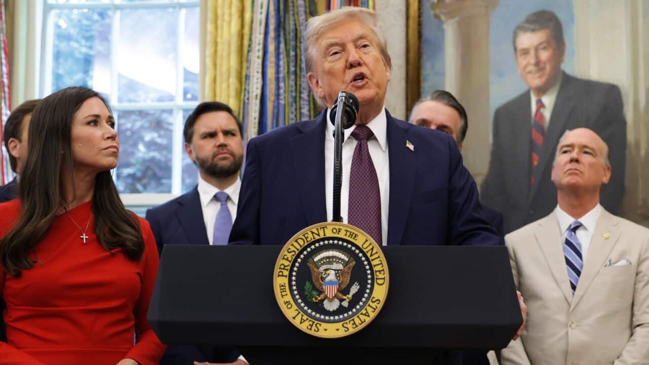 Donald Trump standing and talking from behind a podium inside. A woman in a red dress is standing next to him and behind of him a row of men wearing suits are looking on attentively.