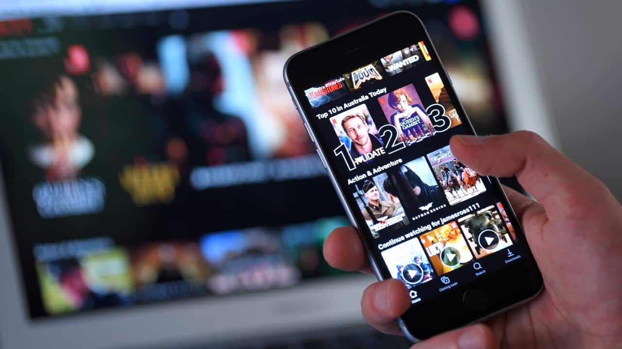 A person holding a phone with the Netflix app open. A laptop in the background also has Netflix open.