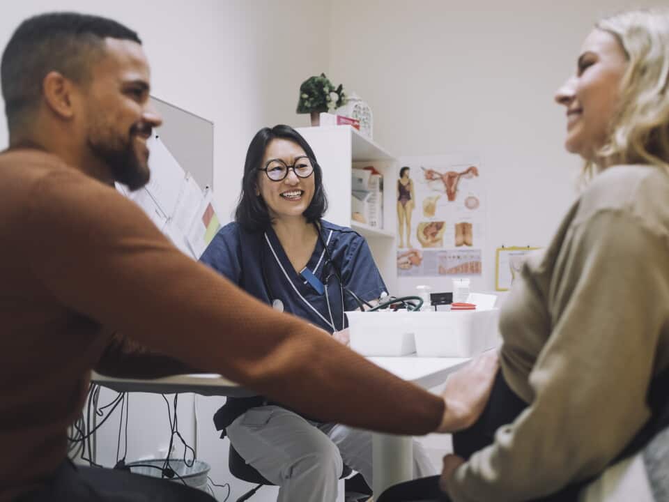 Happy female gynecologist looking at smiling man touching stomach of pregnant woman in clinic