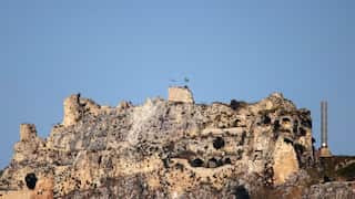 Two flags are flying atop a castle on top of a mountain.