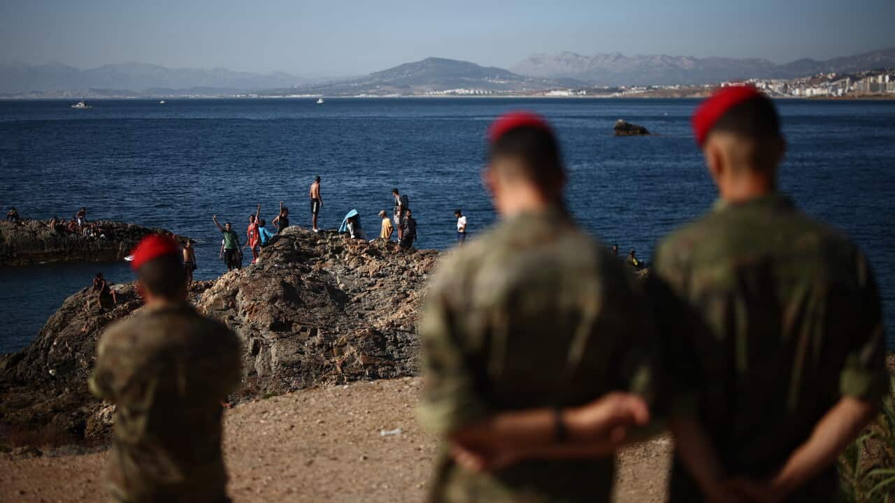 Soldiers look over migrants on a rocky coastline.