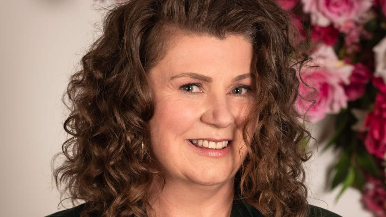 A close-up shot of a middle-aged woman with curly brown hair smiling in front of a background of pink roses.