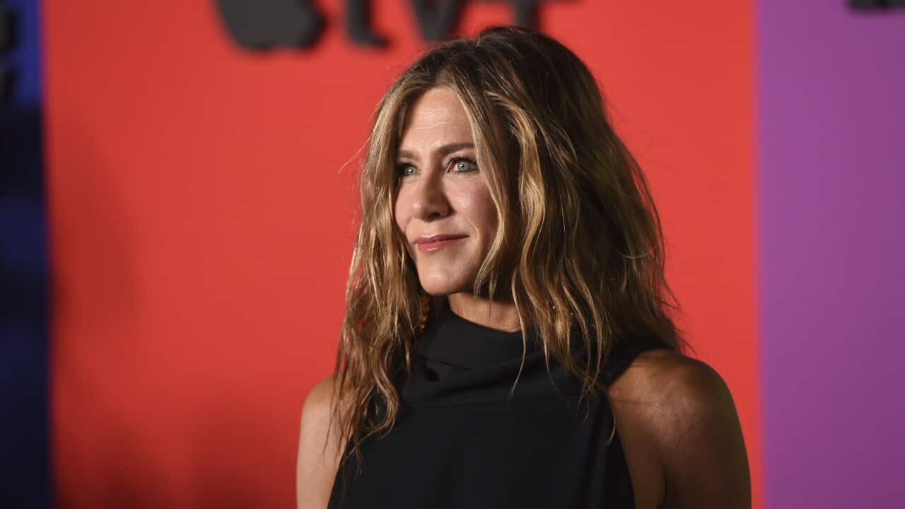 Jennifer Aniston wearing a black dress stands in front of a red screen.