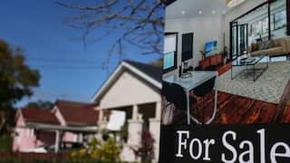 A real estate “For Sale” sign featuring a photo of a home interior, with suburban houses in the background.