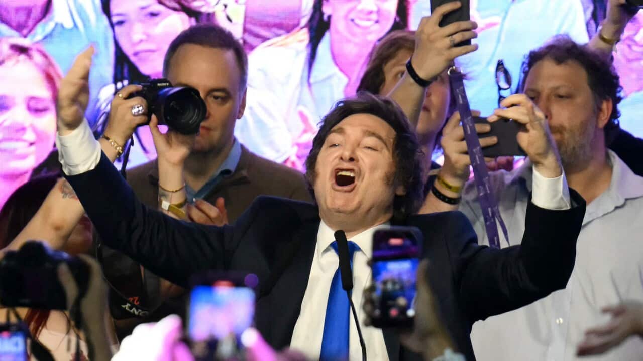 A man in a suit celebrates on a stage with his supporters taking photos of him.