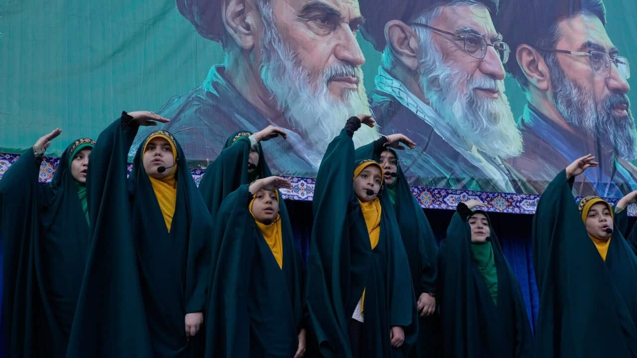 Girls sing as they show the movement of missiles with their hands next to a mural of revolutionary leaders.