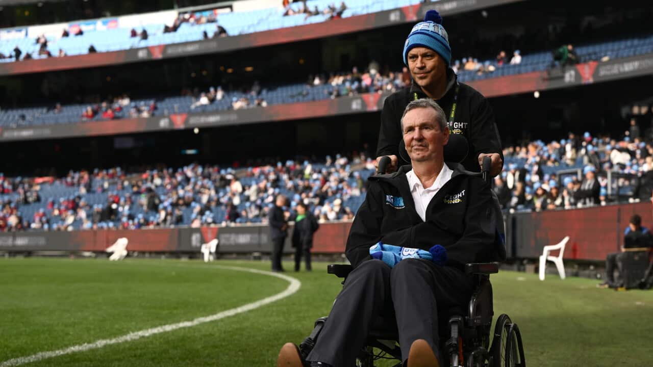 A man in a wheelchair is pushed by a man in a blue beanie on a sports field.