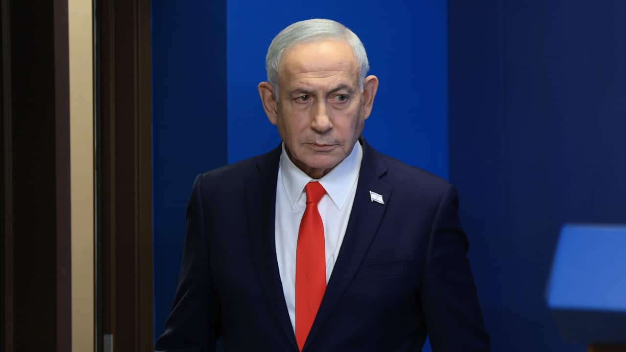 A medium close-up photo shows Israeli Prime Minister Benjamin Netanyahu looking forward with a serious expression, wearing a dark suit, a white shirt, a bright red tie, and an Israeli flag pin on his lapel against a blue background.