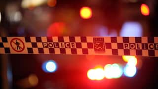 Police tape marked “POLICE” and “Crime Stoppers” stretches across the foreground, with blurred red, blue and amber emergency lights glowing in the background at night.