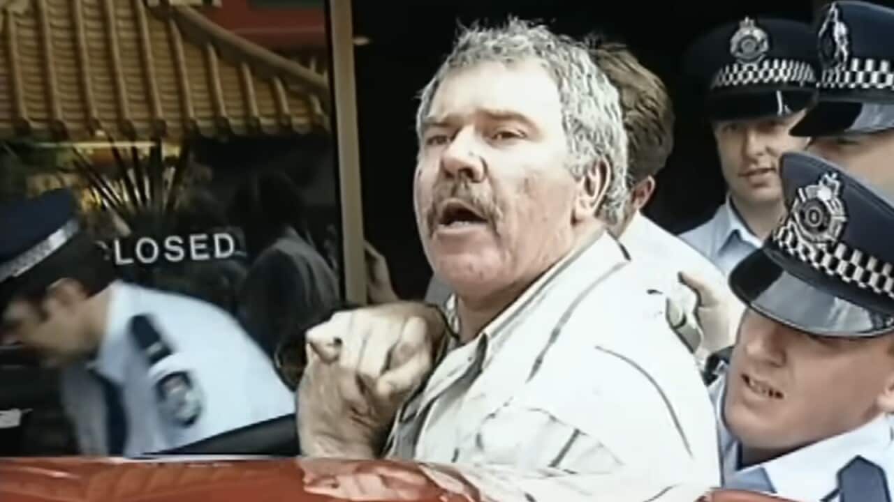 A man with a moustache being arrested by police officers.