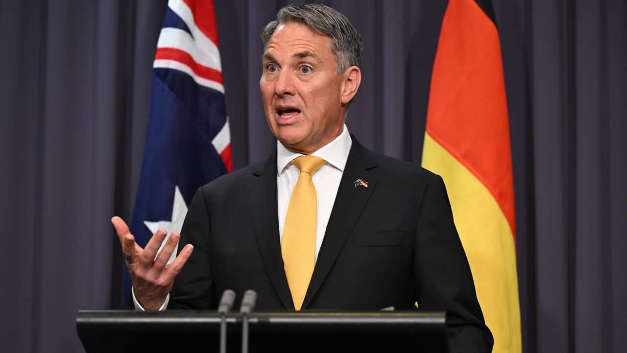A man in a dark suit and yellow tie speaks at a podium, gesturing with one hand.