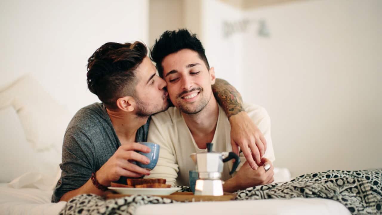 Male couple enjoying breakfast in bed, man kissing partner on cheek