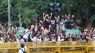 A crowd of demonstrators raise their hands and cheer behind yellow metal barricades near a traffic light and street signs on an outdoor road.