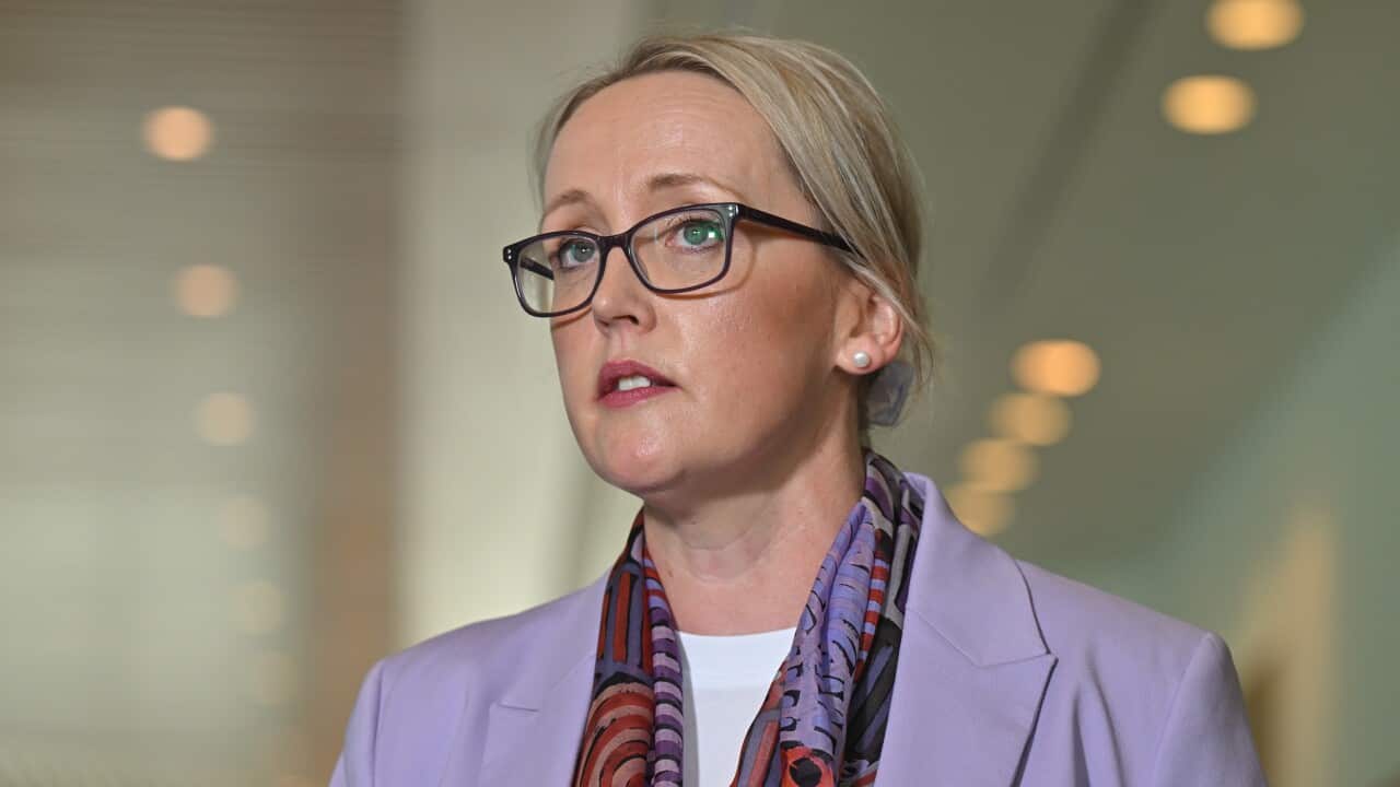 A woman wearing glassess and a purple blazer speaks to reporters.