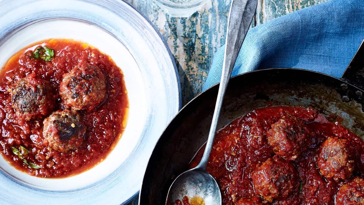 A cast iron saucepan sits on a blue napkin, on a blue-painted wooden surface. The pan is full of meatballs in a rich tomato sauce. A pale blue plate sits beside the pan, with a serve of meatballs. Three slices of fresh bread sit nearby.