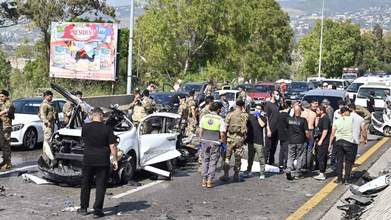 Soldiers and emergency personnel stand around a destroyed white car on a highway
