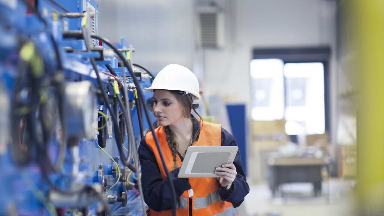 Technician with reflective vest in factory hall inspecting machine with digital tablet