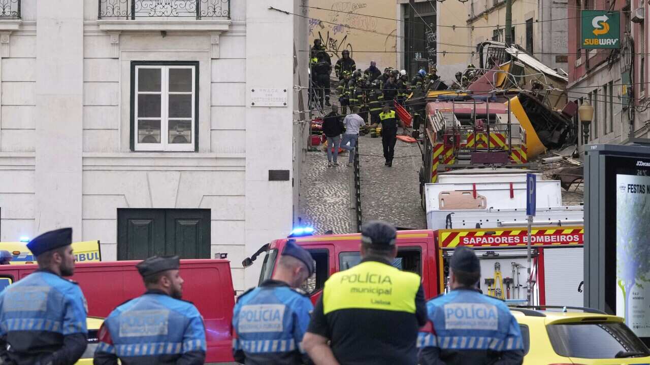 Rear view of a row of five men - four are wearing blue uniforms the the word policia written on the back. There is a fire engine in front of them. The men are looking up a narrow, sloped street at debris. There are also men near the debris.