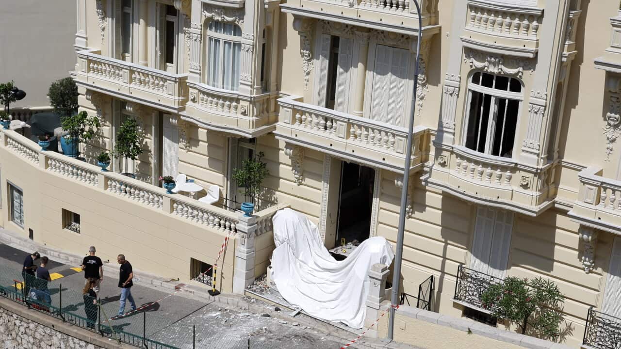 The exterior of a luxury apartment building in Monaco. The building facade is an eggshell white colour. A white sheet is draped across a ground-floor doorway and some debris is spread across the footpath. A small group of people stand near a police cordon nearby.