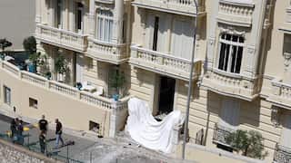 The exterior of a luxury apartment building in Monaco. The building facade is an eggshell white colour. A white sheet is draped across a ground-floor doorway and some debris is spread across the footpath. A small group of people stand near a police cordon nearby.