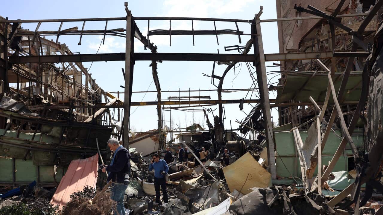 A person with a camera walks through the charred and twisted metal remains of a heavily damaged building surrounded by debris.