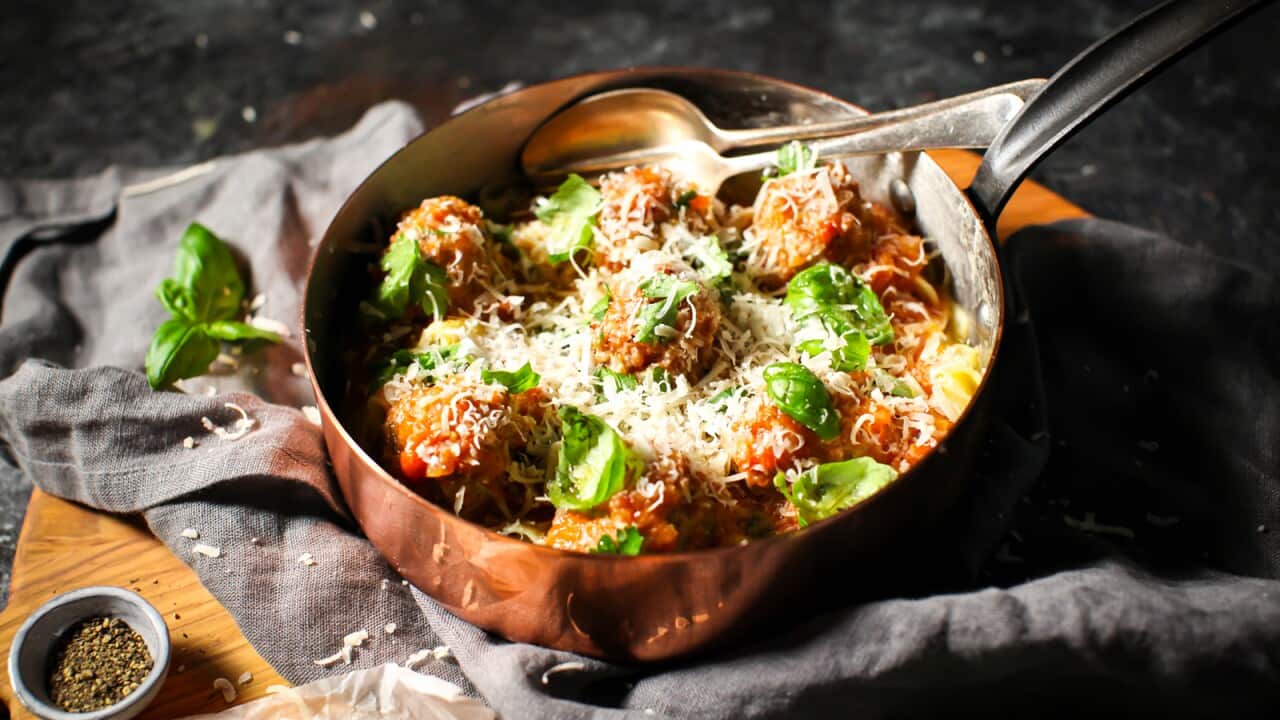 A deep copper saucepan sits on a grey napkin. It holds meatballs and pasta, topped with basil leaves and grated parmesan.