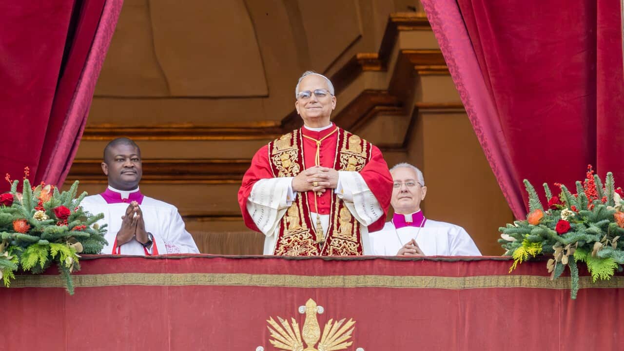 Pope Leo speaking from a balcony at St Peter's Basilica. There are two priests standing on either side of him.