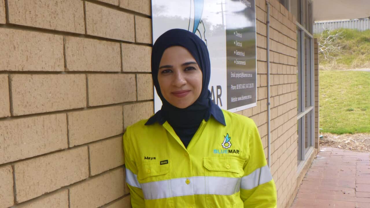 A woman wearing a headscarf and hi-vis clothing leans against a wall
