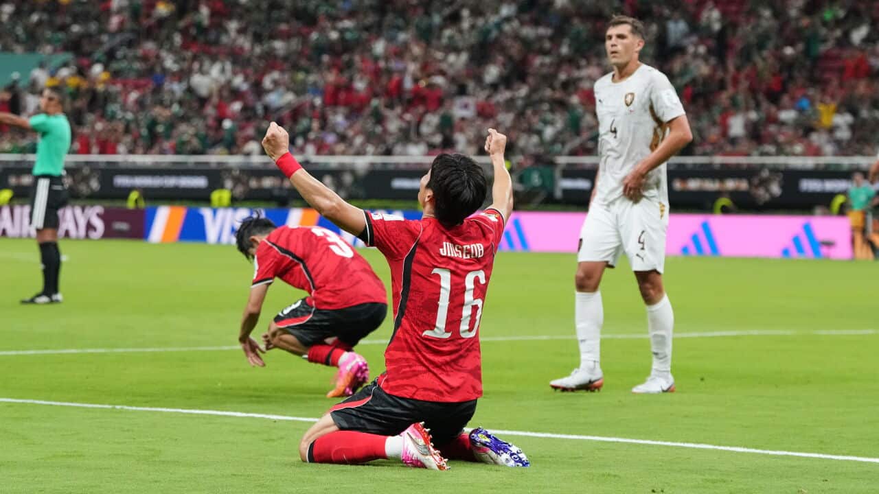 A football player wearing a red jersey celebrates his team's win with his hands up in the air as he kneels on the pitch. Another player wearing a red jersey and another wearing white jerseys are in the background.