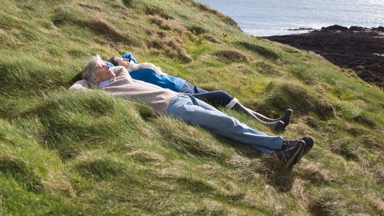 Mature couple lie with hands behind head in coastal landscape