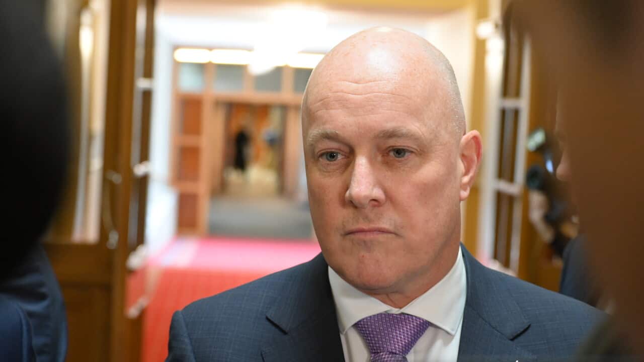 A bald man in a blue suit and purple tie with a blank expression on his face.