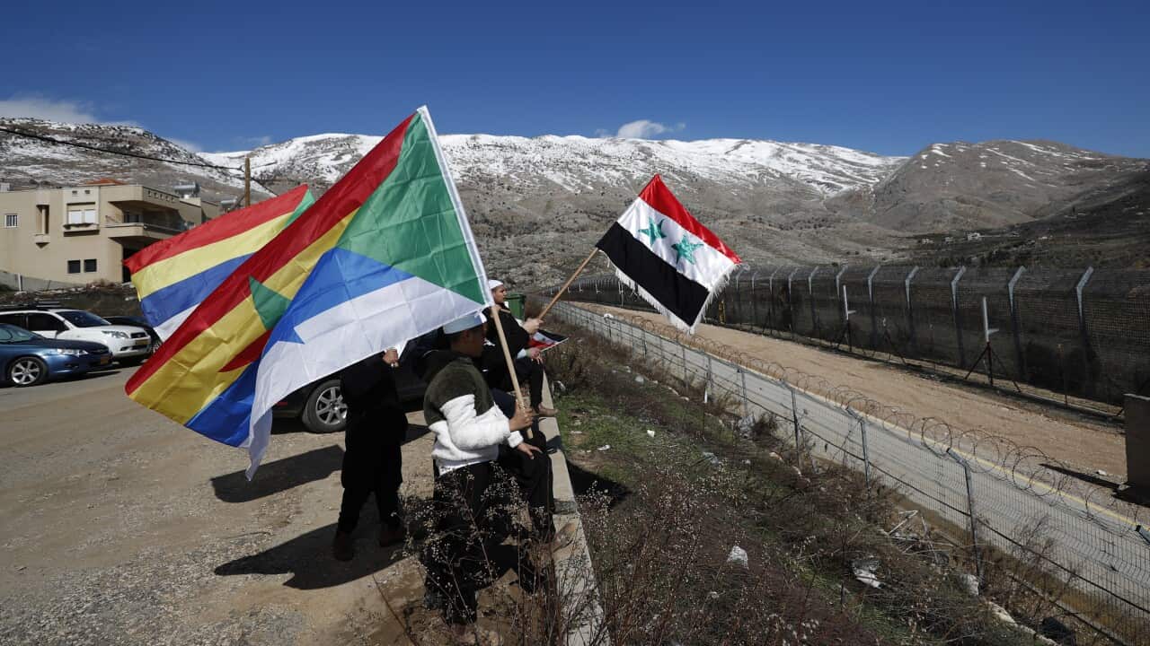 People holding Syrian and Druze flags near a border fence with snow-covered mountains in the background.