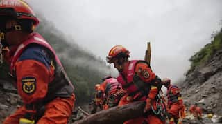 Rescue workers in bright orange uniforms, helmets, and life jackets carry a large log through a muddy landslide area.