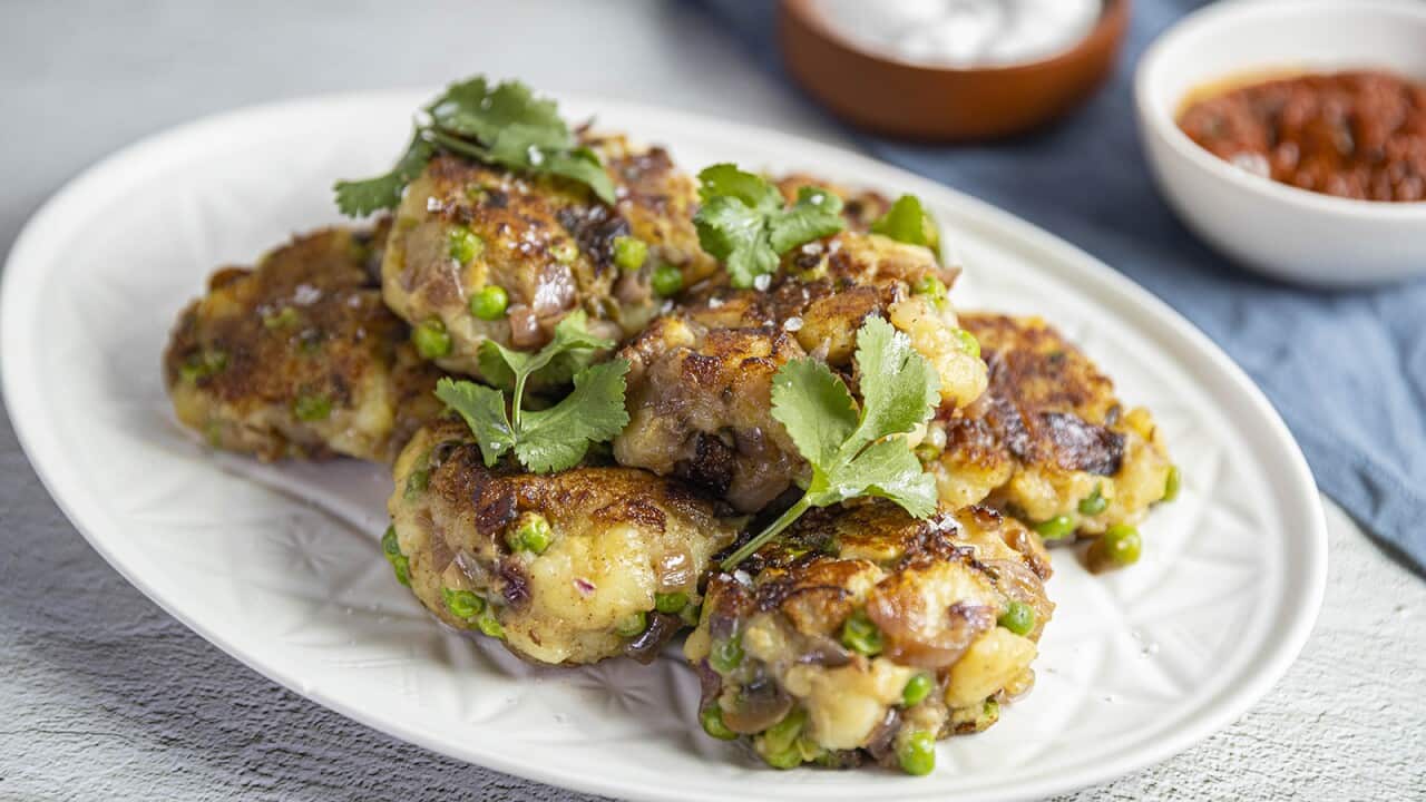 Thick round potato patties, studdied with peas and golden on the top, sit on a textured white plate. Two bowls, one with a red filling and another with a creamy filling, can be seen, out of focus, behind the plate.