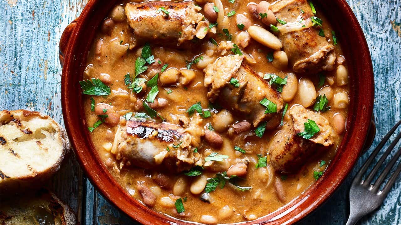 A round brown dish sits on a blue-washed wooden surface. It holds a rustic sausage and bean stew, garnished with a sprinkle of chopped green herbs. Two slices of bread, with grill marks, sit alongside.
