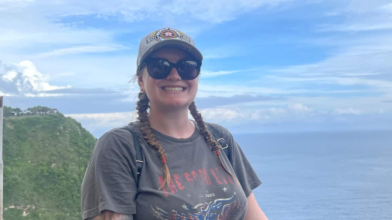 A woman in a grey T-shirt with wings on the front smiles in front of a headland by the ocean